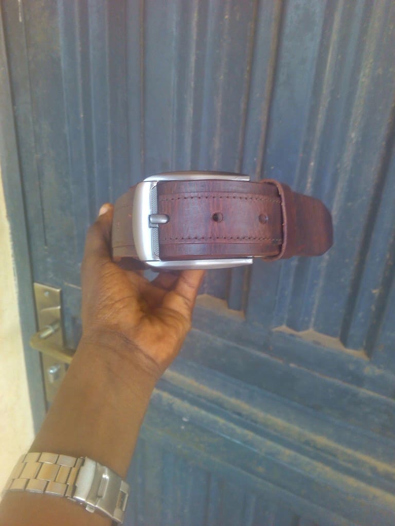 A hand holding up a reddish-brown leather belt with a brushed silver buckle in front of a gray door.