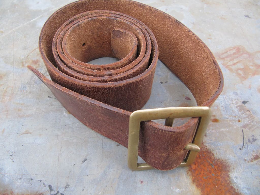 A worn brown leather belt coiled on itself beside a heavy brass buckle, on a weathered concrete surface.