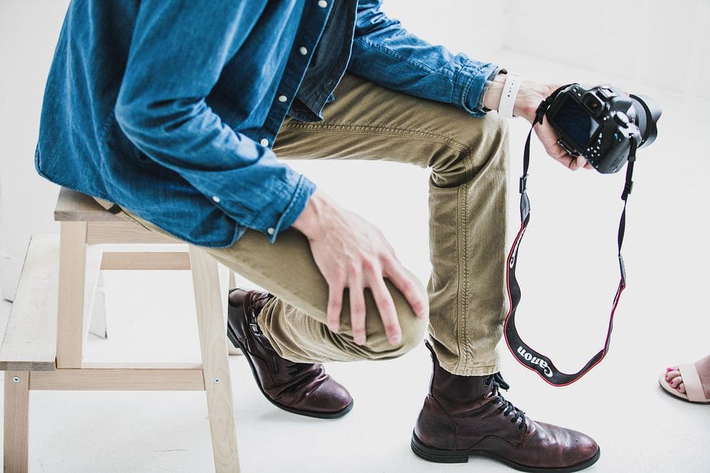 A seated man in slim khaki chinos, a denim shirt and dark brown leather boots, a camera strap in his hand.