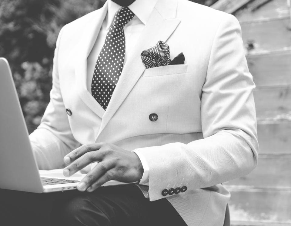 A black-and-white photo of a man in a pale double-breasted jacket, polka-dot tie and pocket square, typing on a laptop on his knee.