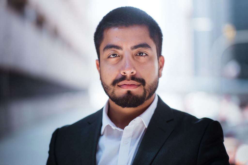A portrait of a bearded man in a dark blazer and open-collar white shirt against a soft, blurred background.