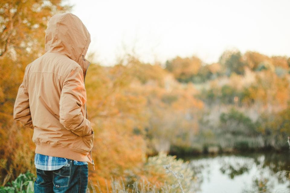 Seen from behind, a man in a tan hooded jacket over a plaid shirt and jeans looks out over a lake ringed with autumn trees.