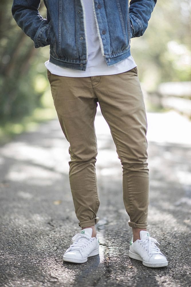 A man in slim khaki chinos with a short, cuffed hem and white sneakers, shot from the waist down on a shady path.