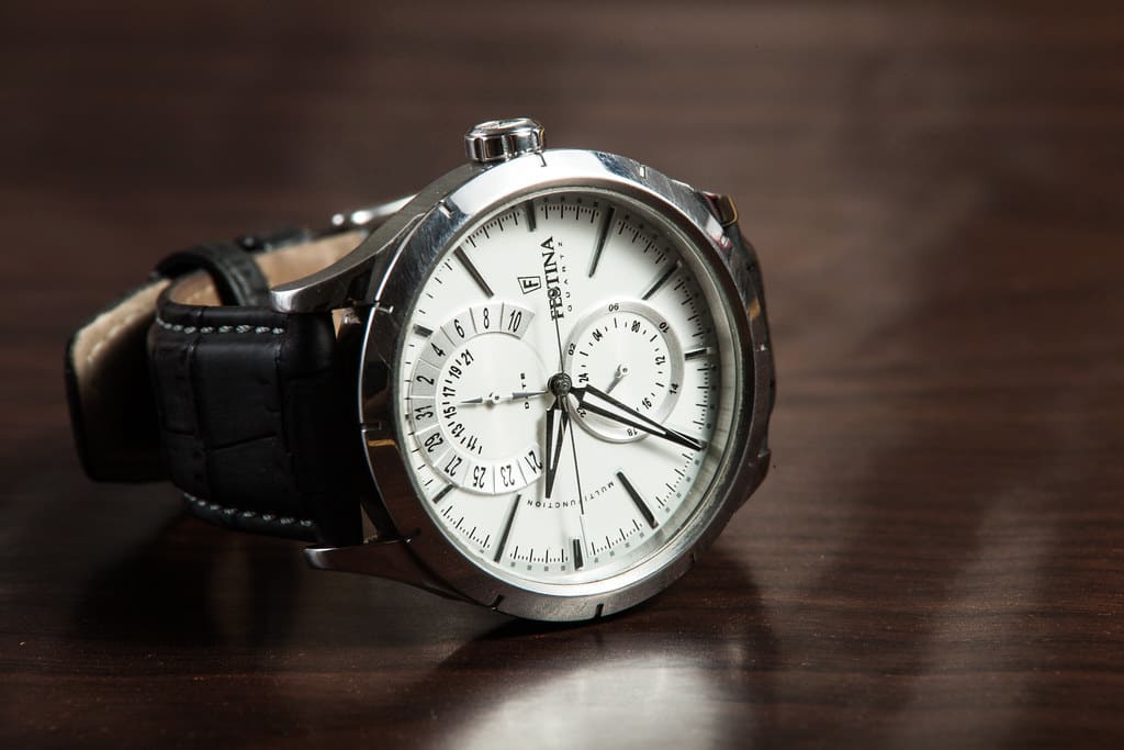 A steel watch with a white dial and a black leather strap lying on a dark wooden surface.