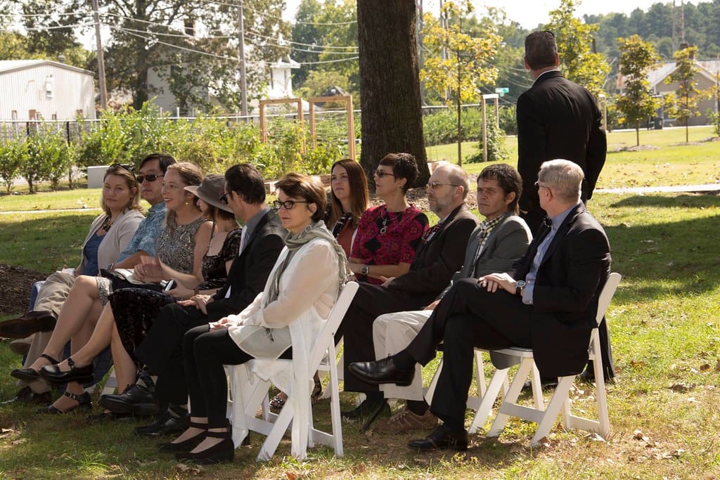 Wedding guests in suits and dresses seated on white folding chairs at an outdoor ceremony under trees.