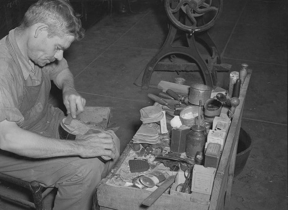 A black-and-white photograph of a cobbler bent over a shoe at his bench, a box of tins, tacks and leather scraps beside him.