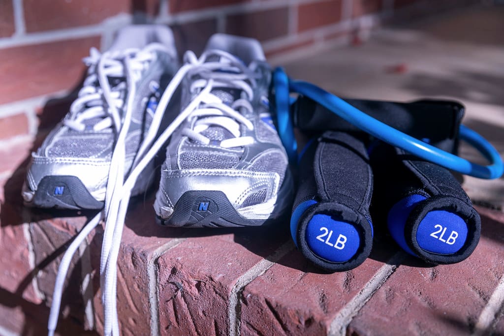 A pair of gray and silver New Balance running shoes on a brick ledge beside blue hand weights.