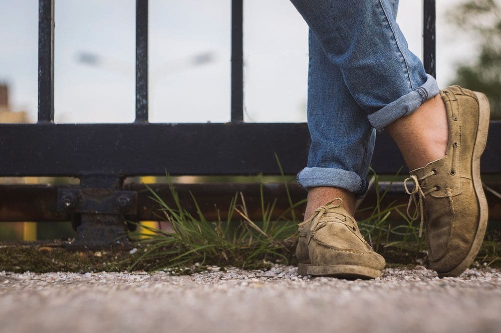 A tan moccasin-style boat shoe worn with cuffed jeans beside an iron fence.